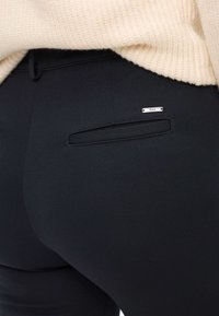 Zwarte nauwsluitende broek van een gestructureerde stof, met een achterzak voorzien van een klein zilverkleurig label. De tailleband is gedeeltelijk zichtbaar onder een lichte trui.