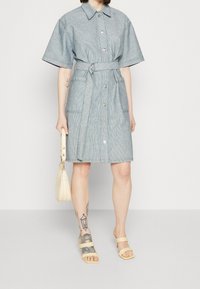 Kvinna i kortärmad grå klänning med bälte, bär en liten beige handväska, beige sandaler, med tatueringar på vänster arm och ben.