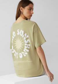 Olijfgroene oversized t-shirt met korte mouwen, voorzien van een grote witte zonafbeelding en tekst op de achterkant, gemaakt van zachte, gestructureerde katoen.