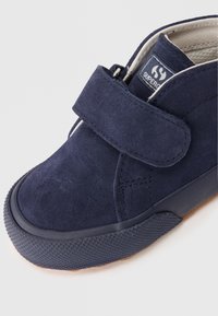 Sneaker in suede blu navy con strap in velcro, punta rotonda, suola in gomma texturizzata e un'etichetta del marchio ben visibile sul lato.