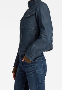 Tmavě modrá denimová bunda s krátkým střihem, vysokým límcem, knoflíky vpředu, dvěma kapsami na hrudi a žebrovanými manžetami.