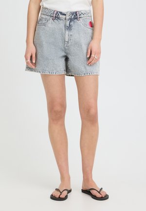 Žena oblečená v svetlosivých rifľových šortkách s ružovým prešívaním a čiernymi žabkami, stojaca pred jednoliatym bielym pozadím.