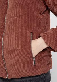 Hnědá kordurová bunda se zapínáním na zip a boční kapsou, s ribkovanou texturou a uvolněným střihem.