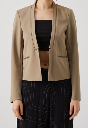 Blazer beige cintré porté ouvert sur un top court noir avec un pantalon noir taille haute comportant de grandes coutures blanches abstraites.