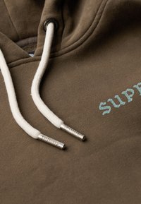 Sudadera marrón con una textura suave, que presenta el logotipo "SUPERDRY" en azul claro y cordones blancos con extremos de metal plateado.
