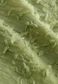 Jasnozielona tkanina o teksturze z postrzępionymi brzegami, ukazująca warstwy i różnice w grubości. Miękki, płynny materiał z delikatnym połyskiem.