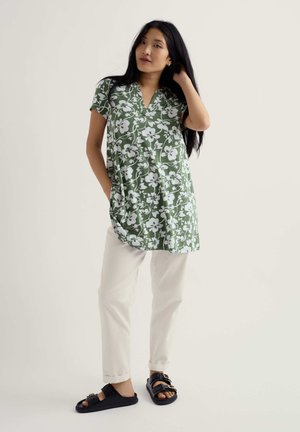 Ženska v zeleni cvetlični tuniki s kratkimi rokavi, belih hlačah in črnih sandalih z zaponko, stoji z eno roko v žepu in drugo dotika las.