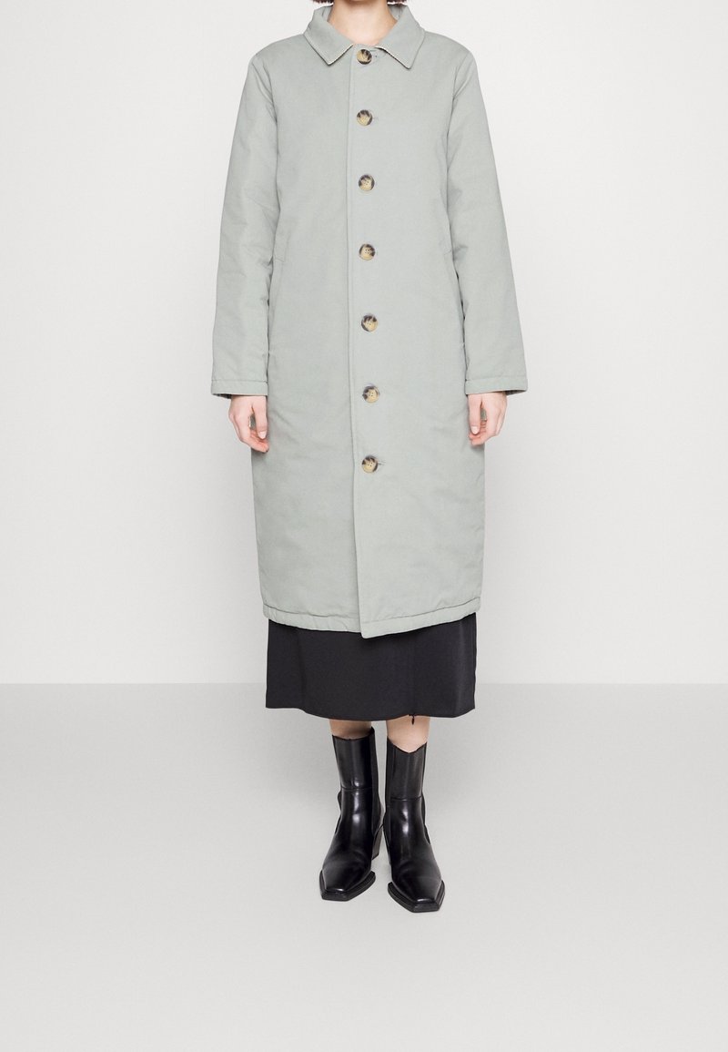 Femme portant un long manteau boutonné gris clair par-dessus une jupe noire et des bottines noires, debout devant un fond blanc uni.