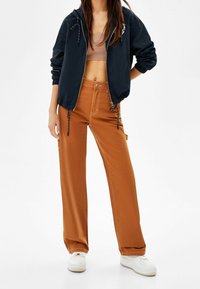 Marinblå lätt vikbar jacka med dragsko i huvan, kombinerad med lösa bruna byxor med sidofickor och kontrasterande sömmar. Vita sneakers.