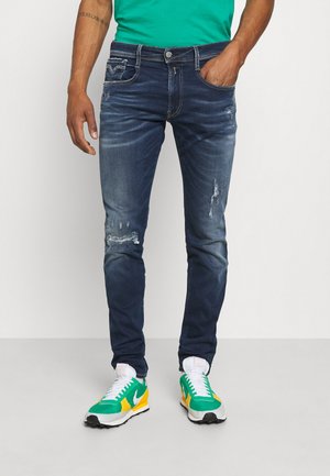 Homme portant un jean slim déchiré bleu foncé avec des baskets Nike colorées vertes, jaunes et blanches, main dans la poche sur fond blanc uni.