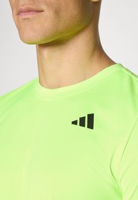 Camiseta deportiva verde fluorescente con cuello redondo, fabricada con un material suave y ligero. Presenta un logotipo geométrico negro en el pecho.