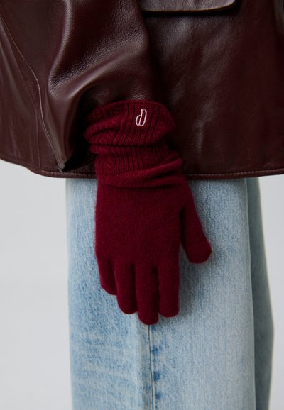 Guantes de punto burdeos con puños acanalados, que presentan aberturas para los dedos y un pequeño logotipo bordado. Chaqueta de cuero sin arrugas al fondo.