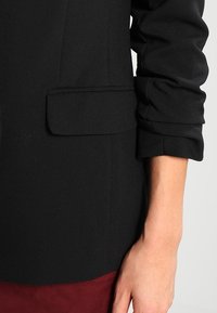 Blazer noir sur mesure avec devant structuré, poche appliquée et détails de manches retroussées, associé à un chemisier texturé bordeaux en dessous.