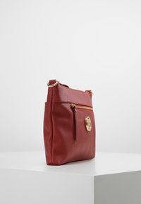 Czerwona torebka crossbody z teksturowanej skóry, z przednią kieszenią na zamek i złotym okrągłym logo, umieszczona na białym piedestale na tle jednolitego tła.