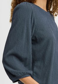 Top blu scuro con texture e maniche a palloncino a tre quarti, caratterizzato da un motivo di tessuto stropicciato e un collo rotondo.