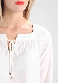 Blouse blanche avec un col rond, des détails cintrés en dessous et une fermeture à nouer à l'avant. Tissu texturé avec un éclat subtil.