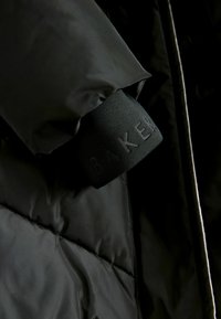 Veste noire rembourrée à la finition lisse et mate. Étiquette de logo portant la mention "BAKER" sur le poignet. Détails texturés et matelassés.