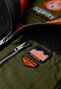 Grøn zip-up jakke med fleecefor, sølvlynlås og flere broderede lapper i orange, blå og hvid.