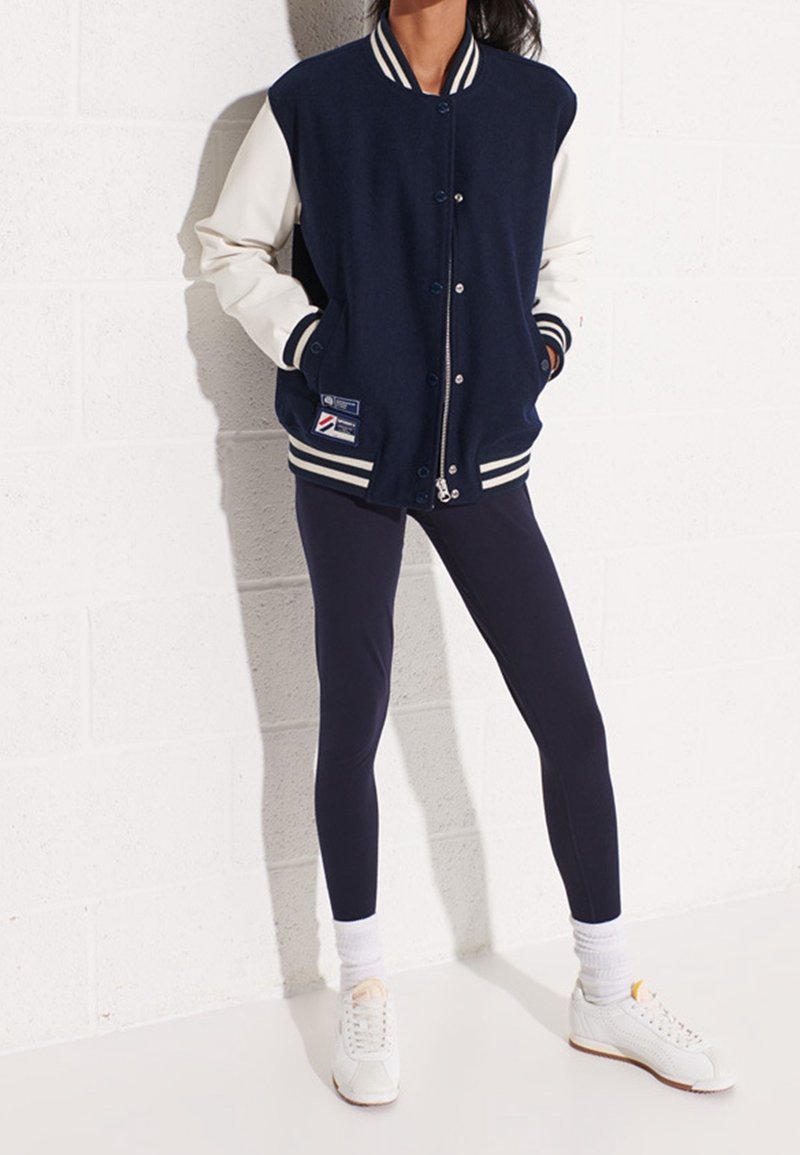Personne portant une veste universitaire bleu marine et blanche, des leggings bleu marine, des chaussettes blanches et des baskets blanches, debout contre un mur blanc.