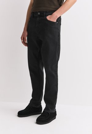 Mann, der schwarze Jeans mit geradem Bein und schwarze Wildlederschuhe trägt, steht mit einer Hand in der Tasche vor einem schlichten weißen Hintergrund.