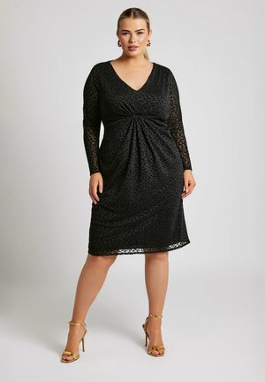 Robe noire avec un décolleté en V, des manches longues en tissu transparent et des fronces à la taille. Présente un subtil motif léopard et une longueur au genou.