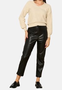 Beiger Strickpullover mit V-Ausschnitt, kombiniert mit hoch geschnittenen schwarzen Lederhosen. Das Modell trägt schwarze Slipper. Textur und Passform betont.