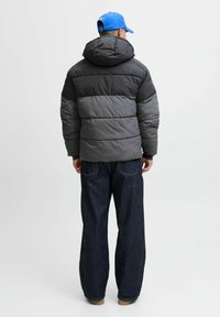 Gepolsterteste Jacke in Grautönen mit Kapuze, mit horizontaler Steppung. Kombiniert mit weit geschnittenen, dunkelblauen Jeans und einer blauen Mütze.