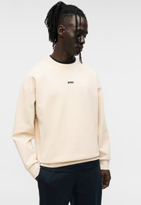 Sweat-shirt beige avec un col rond, épaules tombantes, et logo « BOSS » brodé en noir. Porté avec un pantalon de couleur foncée.