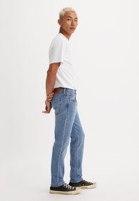 Joven con cabello rubio corto, vistiendo una camiseta blanca, jeans azules y zapatillas negras, de pie de lado con las manos detrás de la espalda.