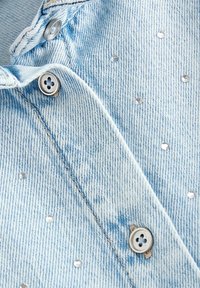 Camicia in denim blu chiaro con bottoni metallici e accenti di piccoli strass trasparenti sparsi. Presenta un colletto classico e un tessuto testurizzato.