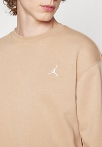 Gros plan sur une personne portant un sweat-shirt beige avec un petit logo blanc de joueur de basketball brodé sur la poitrine.