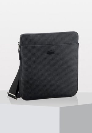 Sac bandoulière noir texturé avec une fine poche avant et une sangle ajustable, exposé sur un piédestal blanc.