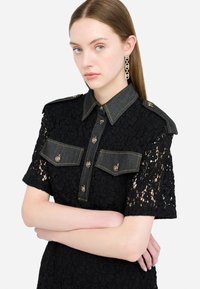 Ung kvinde med glat brunt hår iført sort blonderkjole med denimkrave og lommer, krydsede arme og lange kædeøreringe.