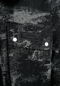 Bolsa de tela lavada en negro y gris con solapa, dos botones de presión de metal blanco, y una etiqueta de cuero negro rectangular cosida en la parte superior.