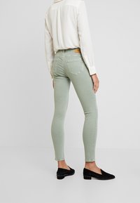 Jeans ajustados en mezcla de algodón verde claro, con cintura alta y dos bolsillos traseros, combinados con una blusa blanca y zapatos planos negros.