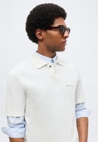 Νέος άνδρας με κοντά σγουρά μαλλιά που φορά γυαλιά ηλίου με πλαίσιο από χέλυδα, λευκό πόλο πάνω από μπλε ριγέ πουκάμισο με τσαλακωμένα μανίκια.