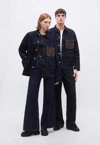 Τζιν μπουφάν ναυτικού μπλε με καφέ τσέπες, συνδυασμένα με τζιν φαρδιάς γραμμής. Λείο υλικό, αντίθετες ραφές και κουμπώματα με πατιλέτες.