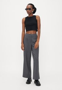 Sort cropped tank top i kombination med grå brede bukser med knaplukning. Modellen bærer sorte solbriller og kraftige sorte sko.