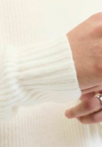 Manga de suéter blanco de punto acanalado con detalle texturizado, tejido suave, y un primer plano de una mano que lleva un anillo de plata.