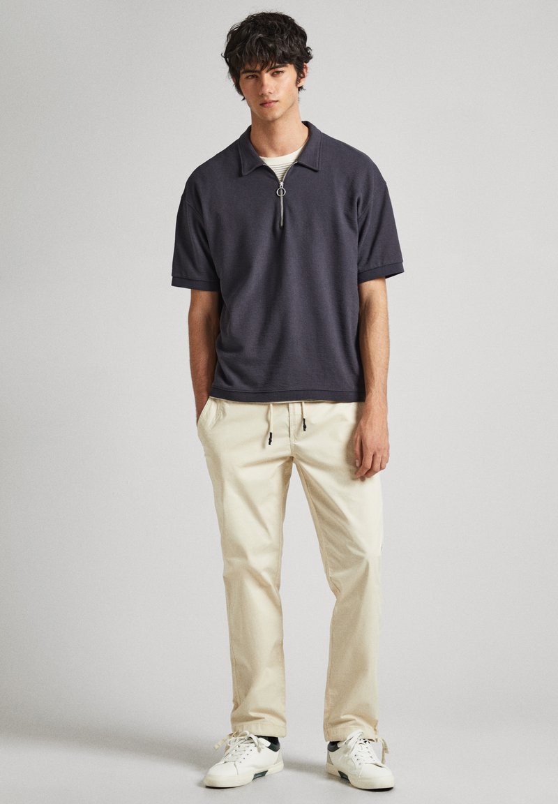 Polo de manga corta gris oscuro con cremallera, combinado con pantalones beige con cordón y zapatillas blancas. Diseño simple, textura suave.