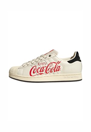 Zapatilla blanca de corte bajo con logo rojo "Enjoy Coca-Cola" en el lateral, lengüeta trasera negra y suela beige con relieve que dice "It's the real thing."