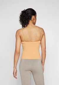 Peegelkaelusega persikka värvi sleeveless topp, mis on voolujooneline, sileda tekstuuriga ja õhukeste rihmadega, koos heledate hallide pahkluu pikkuste leggingsitega.