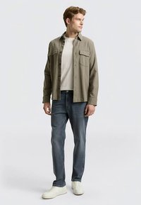 Ljusolivgrön skjorta med knappar, vit T-shirt med rund hals, blå jeans och vita sneakers. Skjortan har två framfickor och en ledig passform.