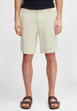 Linned beige shorts med lige snit, forlommer og knaplukning. Båret med brune sandaler med remme på en neutral baggrund.
