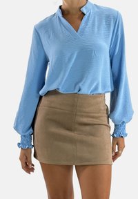 Lichtblauwe blouse met lange mouwen, een V-hals en geplooide blauwe manchetten, gecombineerd met een beige suède minirok met een gladde textuur.
