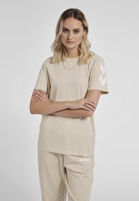 Beige bomulds t-shirt med korte ærmer har hvide chevron-detaljer på venstre arm og et logo på den nedre kant. Løs pasform.