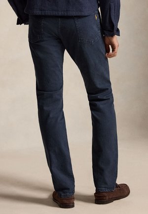 Mężczyzna ubrany w ciemnoniebieskie dżinsy, brązowe mokasyny oraz granatową marynarkę, stojący tyłem na jednolitym beżowym tle.