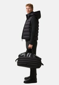 Czarna kamizelka puchowa z kapturem, czarny sweter o fakturze, dopasowane czarne spodnie oraz czarna torba sportowa z błyszczącą górą i uchwytem.