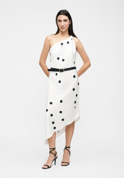 Γυναίκα που φοράει λευκό φόρεμα με ένα ώμο, μαύρες πουά και ασύμμετρο τελείωμα, αξεσουάρ με μαύρη ζώνη και ψηλοτάκουνα πέδιλα με λουράκια.