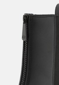 Stivaletto nero con una parte superiore in sintetico liscio, lati elasticizzati e una cerniera metallica con un tirante con marchio.